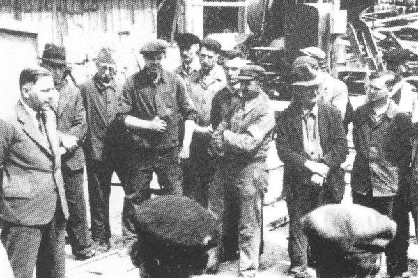 Ein altes Schwarz-Weiß-Foto von der Historie von Brömer & Sohn. Ein Mann in einem Anzug hält eine Ansprache vor einer Gruppe von Arbeitern. Im Hintergrund ist eine Baumaschine.
