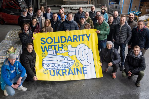 Das Team von Brömer steht zusammen und hält ein gelbes Plakat, auf dem geschrieben steht „Solidarity with Ukraine“. Darauf abgebildet ist eine Hand, die mit zwei Fingern ein Panzerrohr schließt.