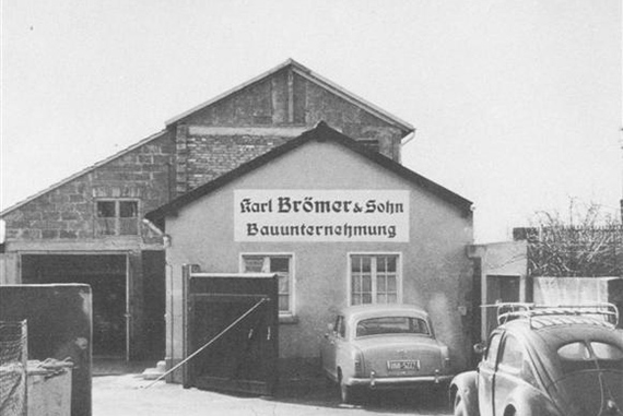 Ein altes Schwarz-Weiß-Foto vom Unternehmenssitz von Brömer & Sohn. Es handelt sich um ein kleines Haus mit Anbau und zwei Autos im Vordergrund.